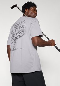 Svetlo siva majica s kratkimi rokavi, ki na hrbtni strani prikazuje grafiko drevesa in golfista, z besedilom "Nike Golf" nad njim in "Samo storite to." spodaj.