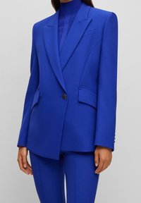 Dopasowana niebieska marynarka i pasujące spodnie wykonane z gładkiej tkaniny. Charakteryzują się wyraźnymi klapami, dwoma przednimi kieszeniami oraz pojedynczym zapięciem na guzik.