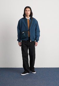 Chaqueta bomber azul con cremallera frontal, suéter marrón debajo, pantalones negros y zapatillas deportivas negras. Incluye una bolsa cruzada de tela en color azul verde.