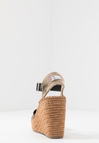 Klínová sandála s patou z pletené juty, béžovým látkovým páskem kolem kotníku, černými akcenty a texturovaným svrškem s přezkou.