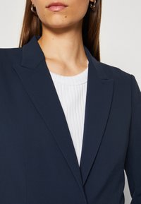 Laivastonsininen bleiseri terävästi leikatuilla läpillä, rakenteellisella istuvuudella ja yhdellä napilla, päällä valkoinen röyhelötoppi. Sujuva kangas ja siistit linjat.