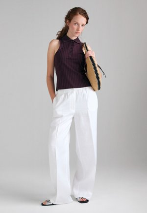 Femme portant un haut sans manches violet foncé et un pantalon large blanc, tenant un sac tissé sur l’épaule, debout devant un fond gris uni.