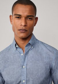Camisa de manga larga con un patrón floral azul, cuello con botones y botones blancos. Tela de algodón suave con un diseño ajustado.