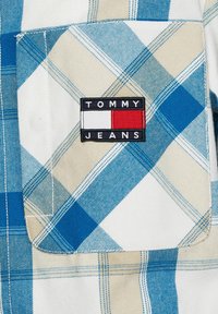 Blå och beige rutig kortärmad skjorta med bröstficka, med en svart etikett som säger "TOMMY JEANS" med röd, vit och blå accent.