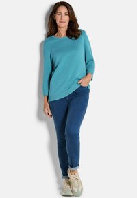 Frau, die an der Wand lehnt und einen strukturierten türkisfarbenen Pullover, dunkle enge Jeans und beigefarbene Sneakers mit Schnürsenkeln trägt.