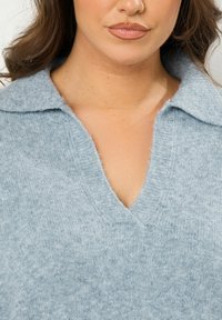 Helles blaues Strickpullover mit einem V-Ausschnitt und einem Kragen. Weiche Textur mit sichtbaren Nähdetails und lässiger Passform.