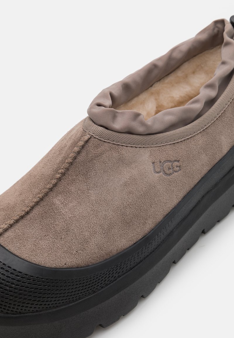 Braune Wildleder-Slipper mit weicher Innenausstattung, elastischem Kragen, schwarzer Gummisohle und subtiler Logo-Detailierung an der Seite.