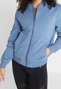 Kvinna som bär en blå bomberjacka med dragkedja, med händerna i fickorna, och svart leggings med nätdetaljer och texten "AIR".