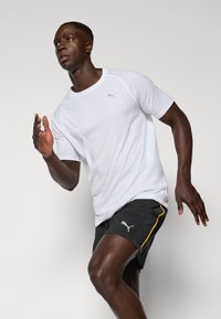 Camiseta deportiva blanca de manga corta con detalles en las costuras, combinada con pantalones cortos negros que tienen una franja lateral amarilla y un logo.
