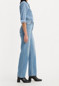 Jeans in denim blu chiaro con vita alta e gambe larghe, abbinati a una camicia a maniche lunghe blu chiaro con bottoni e stivali neri con tacco.