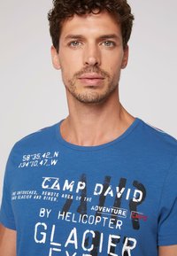 Camiseta de algodón azul con cuello redondo, con texto impreso en blanco y negro, y detalles en rojo. Textura suave y corte casual.