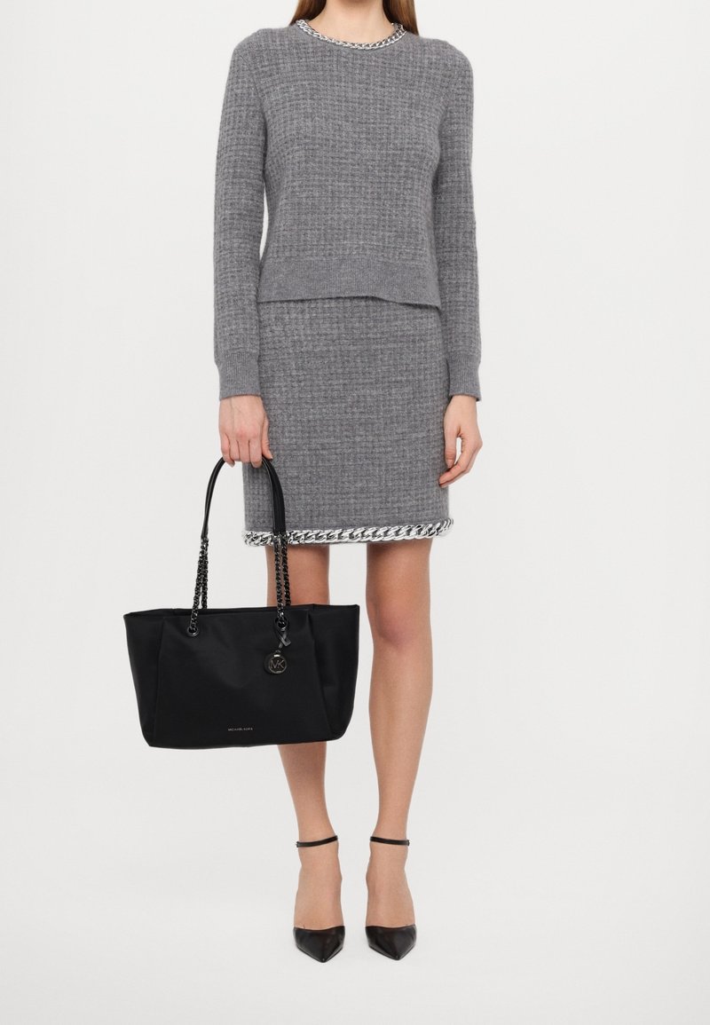 Femme portant un pull en maille gris et une jupe assortie avec bordure en chaîne, tenant un sac à main noir, portant des talons noirs à bride à la cheville.