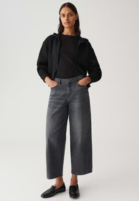 Felpa nera con zip sopra un top nero a coste, abbinata a jeans grigi scuri a vita alta e gamba larga, e scarpe nere slip-on. Sfondo semplice.