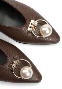 Brązowe płaskie buty ze spiczastym noskiem, ozdobione złotym okrągłym elementem i dużymi białymi perłami na białym tle.