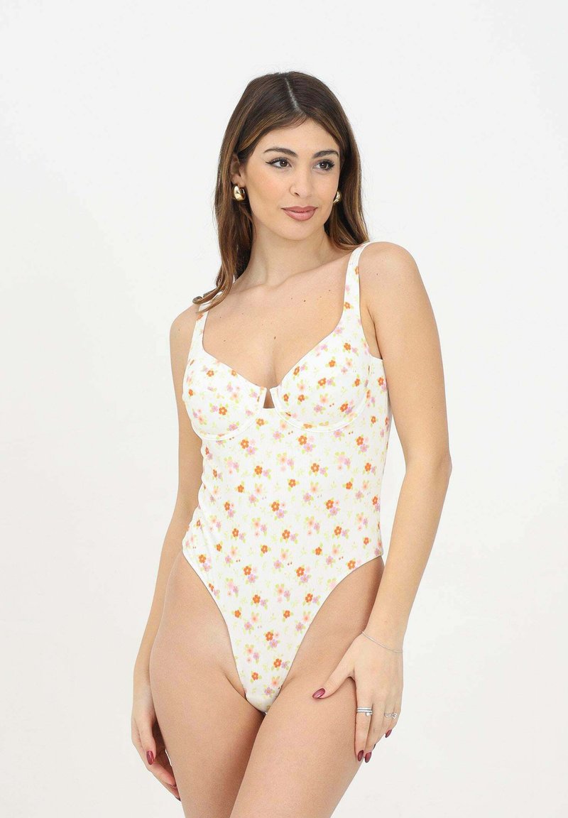 Costume da bagno intero bianco con piccolo motivo floreale in arancione e rosa. Presenta spalline regolabili e un dettaglio a scollatura a U sul busto.