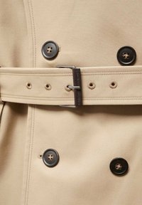 Beige trenchcoat stof met dubbele rij knopen, voorzien van donkere knopen, een bijpassende riem en zichtbare stikseldetails.