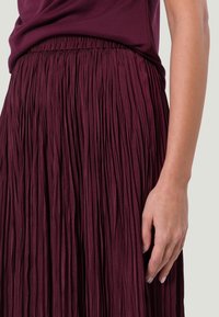 Gonna plissettata bordeaux con vita elastica; tessuto testurizzato e fluente che crea pieghe verticali; abbinata a un top coordinato.