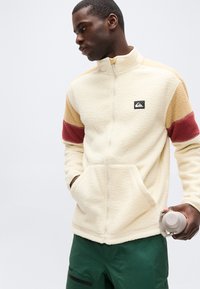 Άνδρας που φοράει μπεζ φλις μπουφάν με κόκκινες και καφέ ρίγες στα μανίκια, πράσινο παντελόνι, κρατώντας ένα γκρι παγούρι στο δεξί του χέρι.