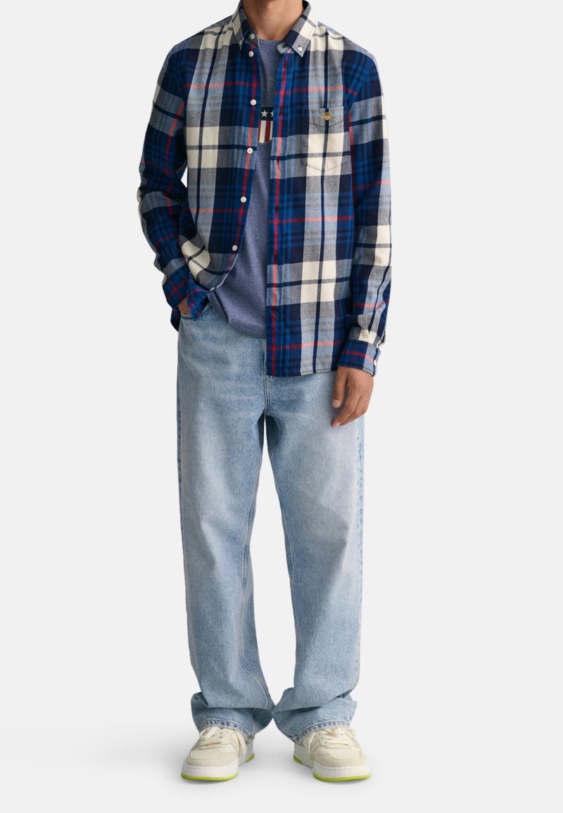 Homme portant une chemise à carreaux bleue, blanche et rouge par-dessus un t-shirt graphique bleu, un jean bleu clair et des baskets blanches avec des semelles vert fluo.