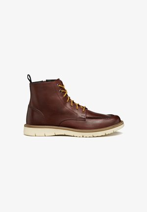 Bottines en cuir marron avec des lacets jaunes, bout rond, semelle en caoutchouc beige et une languette noire à l'arrière. Texture lisse avec un minimum de détails.