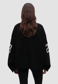 Übergroßer schwarzer Pullover mit gerippter Textur, fransigem Saum und kontrastierender weißer Zahl "23" am Ärmel.