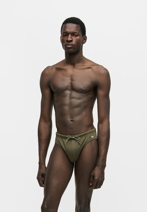 Homme athlétique debout, portant un slip de bain vert olive avec un cordon, sur un fond uni gris clair.