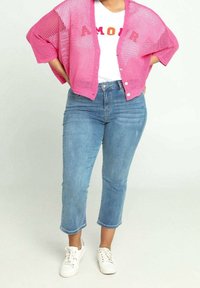 Rosa öppenvävd kofta över en vit t-shirt med röd och blå text, tillsammans med ljusblå cropped jeans och vita sneakers.