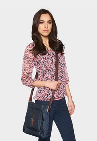 Schoudertas van marineblauw stof met een voorzak, bruine accenten en een verstelbare schouderriem; gecombineerd met een gebloemde blouse in roze en rood.