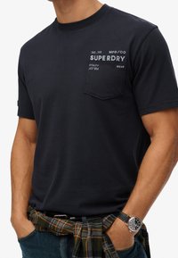 Marineblå bomulds t-shirt med korte ærmer, rund halsudskæring og en brystlomme med hvidt trykt tekst. Parret med en ternet flannel, knyttet om taljen.