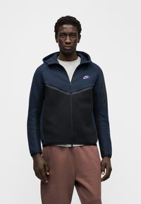 Tumši zils un melns kapučjakas džemperis ar rāvējslēdzēju un chevrona dizainu, uz kura redzams violets logotips un sānu rāvējslēdzēja kabata. Izgatavots no mīksta, teksturēta auduma.