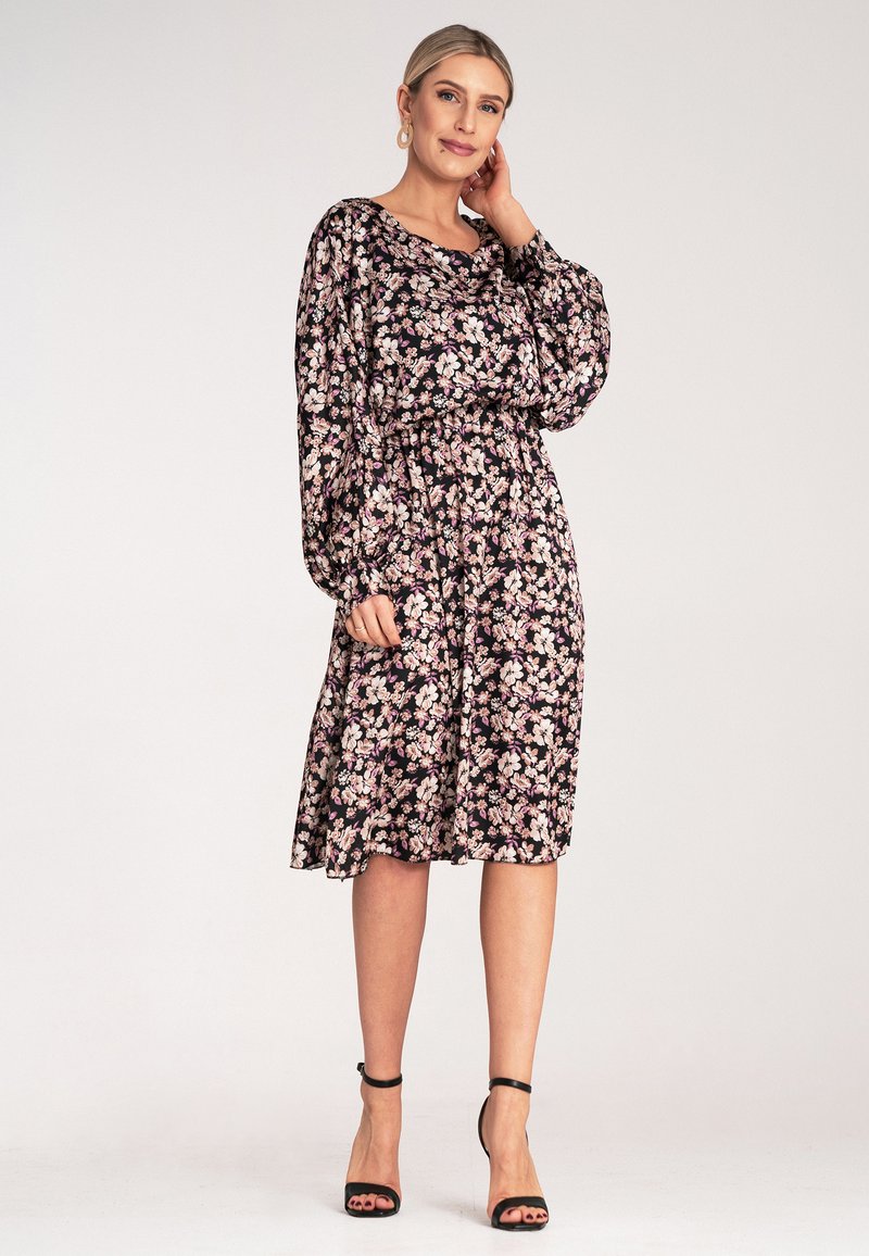 Kwiatowa sukienka w czerni z różowymi i białymi kwiatami, długie rękawy, elastyczny pas i długość do kolan. Połączona z czarnymi sandałami na obcasie.