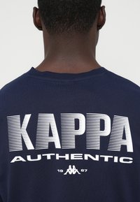 Person som bär en marinblå skjorta med vit text "KAPPA AUTHENTIC 1967" och logotypen av två ryggen mot ryggen sittande figurer på baksidan.
