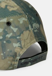 Gorra de camuflaje en tonos de verde y marrón claro, hecha de tejido texturizado. Presenta orificios de ventilación y una correa negra de marca en la parte posterior.