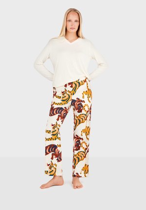 Woman standing barefoot wearing a long-sleeve white top and white pants with multicolored tiger print on a plain light background.