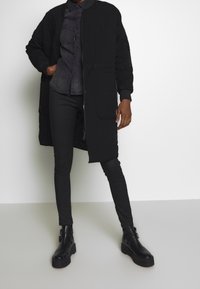 Svart kviltad jacka med lätt lyster, långa ärmar, sidofickor och dragkedja. Bärs över en mörk skjorta med knappar och smala svarta byxor.