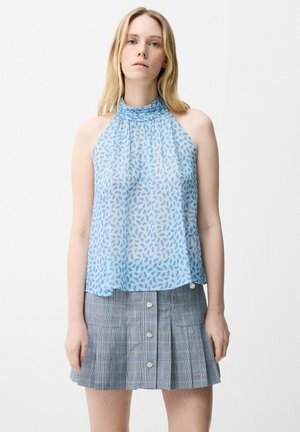 Femme aux longs cheveux blonds portant un haut sans manches bleu clair à motifs et une jupe plissée grise avec boutons, se tenant devant un fond blanc.