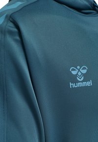 Felpa sportiva color teal con tessuto testurizzato, strisce azzurro chiaro a contrasto e un logo "hummel" ben visibile nella zona del petto.