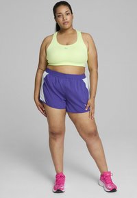 Svetlozelena športna modrčka, vijolične hlače s belimi detajli in rožnate športne čevlje. Osredotočeno na udobje in gibljivost v dizajnu aktivne obleke.