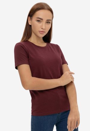 Giovane donna con capelli castani lisci che indossa una maglietta a maniche corte color granata e jeans blu, che si sistema la manica sinistra.