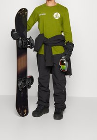 Maglietta verde a maniche lunghe, pantaloni da neve neri e guanti, snowboard nero con attacchi e occhiali riflettenti, tutti adatti per sport invernali.