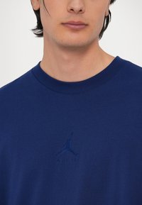 Námořní modré bavlněné tričko s kulatým výstřihem, které má na hrudi embosované logo Air Jordan, hladkou texturu a krátké rukávy.
