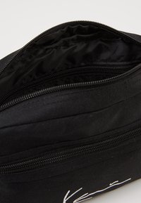 Schwarze Stofftasche mit einem Reißverschluss, die ein glattes Innere und weiße Textdetails auf der Außenseite aufweist. Texturierte Oberfläche.