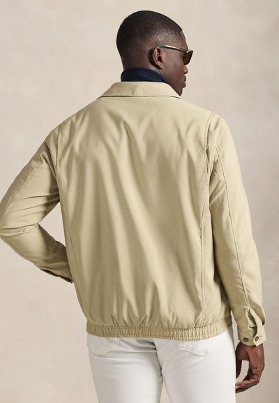 Casaco leve bege com gola alta, bainha elástica e mangas compridas. Textura suave, design minimalista e detalhes sutis de costura.