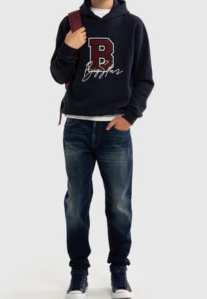 Osoba ubrana w granatową bluzę z kapturem z dużym logo „B Big Star”, niebieskie dżinsy, trampki, nosząca bordowy pasek plecaka na jednym ramieniu.