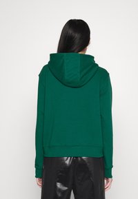 Felpa verde con cappuccio e vestibilità rilassata, caratterizzata da un grande cappuccio e polsini a coste, abbinata a pantaloni neri lucidi a vita alta.