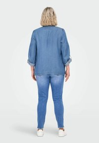 Chaqueta de mezclilla azul claro con mangas arremangadas, combinada con jeans ajustados azul claro. Modelo de espaldas, mostrando un ajuste casual. Zapatillas blancas.