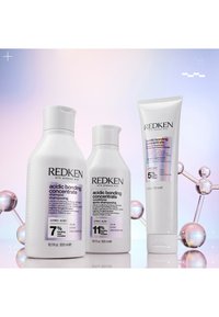 Drei Redken-Haarpflegeprodukte: eine weiße Shampoo-Flasche, eine weiße Conditioner-Flasche und eine weiße Tube mit Leave-in-Behandlung, die mit lila Text versehen sind.
