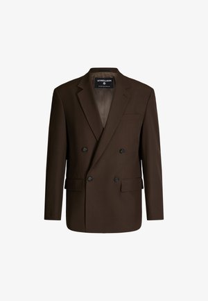 Veste marron croisée à boutons noirs, avec des revers structurés et une poche poitrine. Tissu lisse et doublure texturée visible.