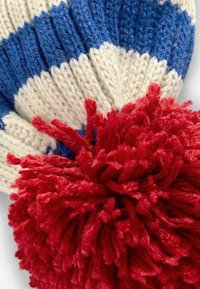 Bonnet en tricot avec des rayures bleues et crèmes, surmonté d'un grand pompon rouge duveteux. Le matériau semble doux et texturé.