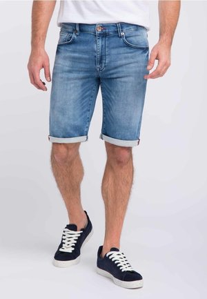 Pantalones cortos de denim en azul claro con un acabado desgastado, con puños enrollados y cinco bolsillos, combinados con zapatillas de ante azul marino con cordones blancos.